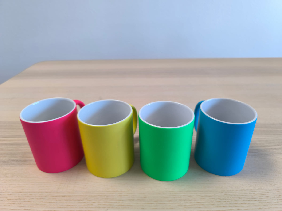 Vier Neon-Tassen in Pink, Gelb, Grün und Blau nebeneinander auf einem Holztisch